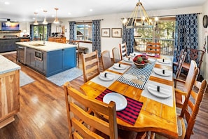 A modern kitchen with a central island, wooden dining table set for four, and a view of the outdoors through large windows.