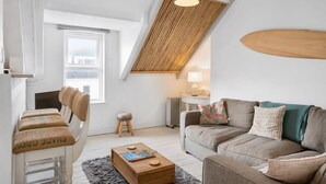 A cozy living room with a high ceiling, a wooden slat ceiling, a sofa, a coffee table, and a chair with cushions. - Beach-Style Flat, 2 Min Walk to Tolcarne Beach (Newquay)