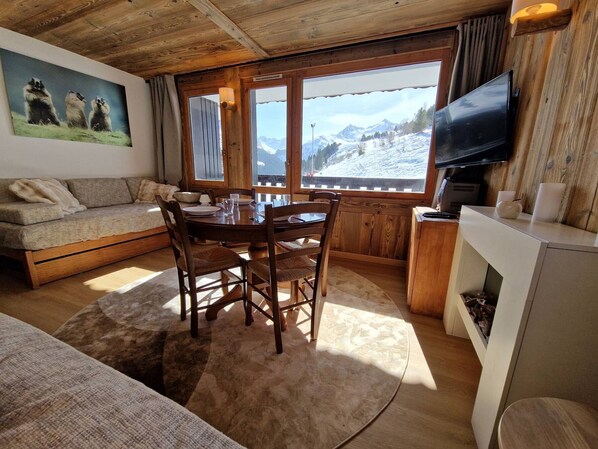 Una habitación acogedora con techo de madera, una mesa de comedor con sillas, un sofá, un televisor y una ventana con vistas a la montaña.