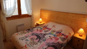 A bedroom with a wooden headboard, two bedside tables with lamps, a window with sheer curtains, and a bed with a colorful Paris-themed bedspread.