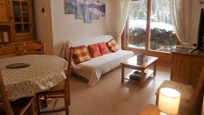 A cozy living room with a sofa, a dining table, and a TV. There are paintings on the wall and a window with curtains.