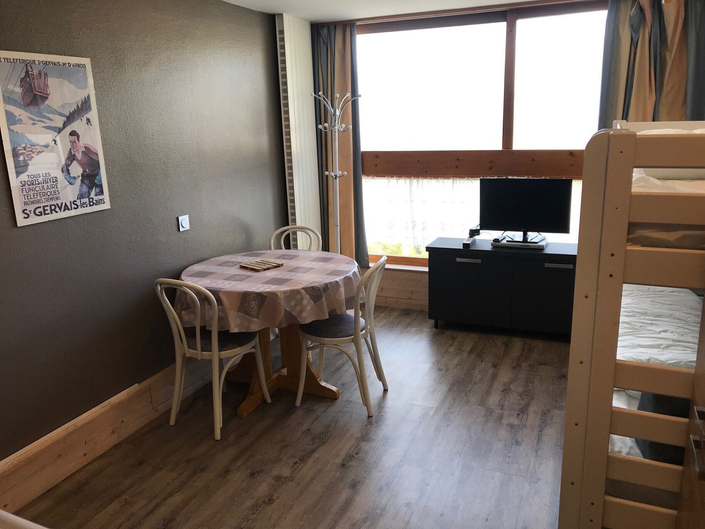 A room with a round table, two chairs, a bunk bed, a TV, and a poster on the wall.