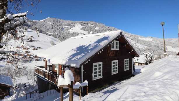 Una cabaña de madera con techo nevado, rodeada de un paisaje invernal y montañas.