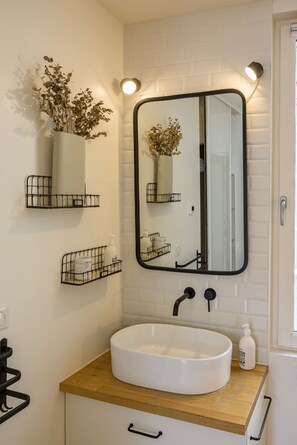 Een badkamer met een witte wasbak, een zwarte kraan en een spiegel met een zwarte lijst. Twee wandplanken met gedroogde planten en toiletartikelen.