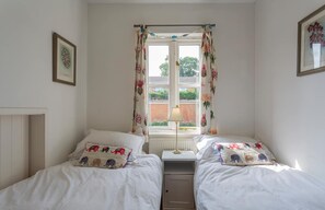 A bedroom with two single beds, a window with floral curtains, a bedside table with a lamp, and a radiator.