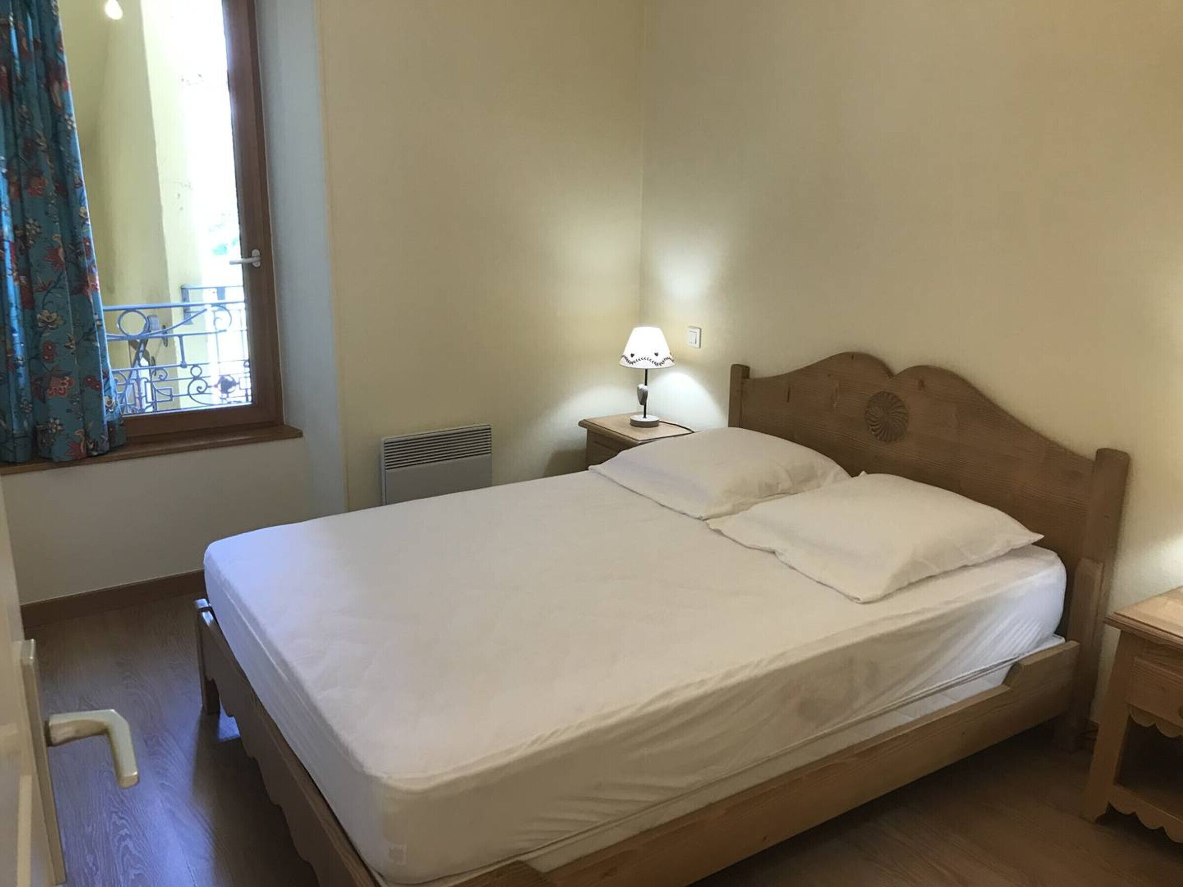 A single bed with a wooden headboard, a bedside table with a lamp, a radiator, and a window with curtains.