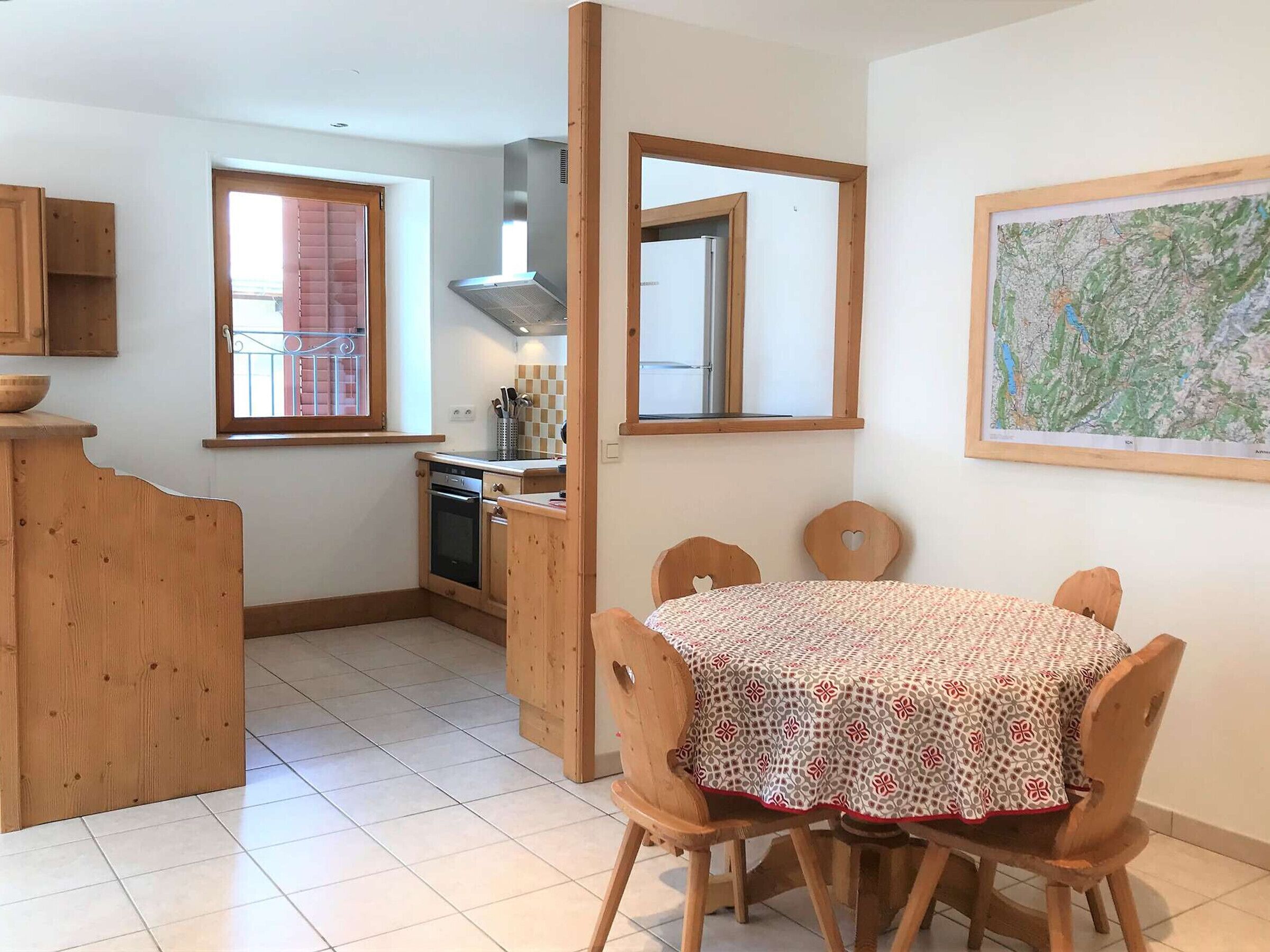 A compact kitchen with wooden cabinets, a dining table with four chairs, and a large window with blinds.