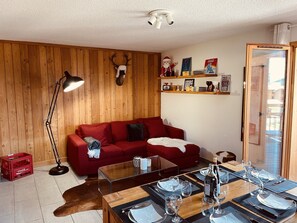 A cozy living room with a red sofa, wooden walls, a glass coffee table, and a dining area set for four.