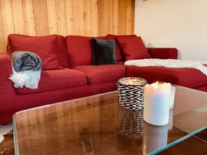 A red sofa with a grey and white throw blanket, a glass coffee table with two candles, and a patterned candle holder.