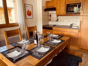 A dining area with a wooden table set for four, a bottle of wine, and wine glasses. There is a kitchen with wooden cabinets and a microwave above the sink.