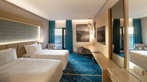 Quarto de hotel com duas camas, uma escrivaninha, uma cadeira, um espelho grande e vista para o mar.
