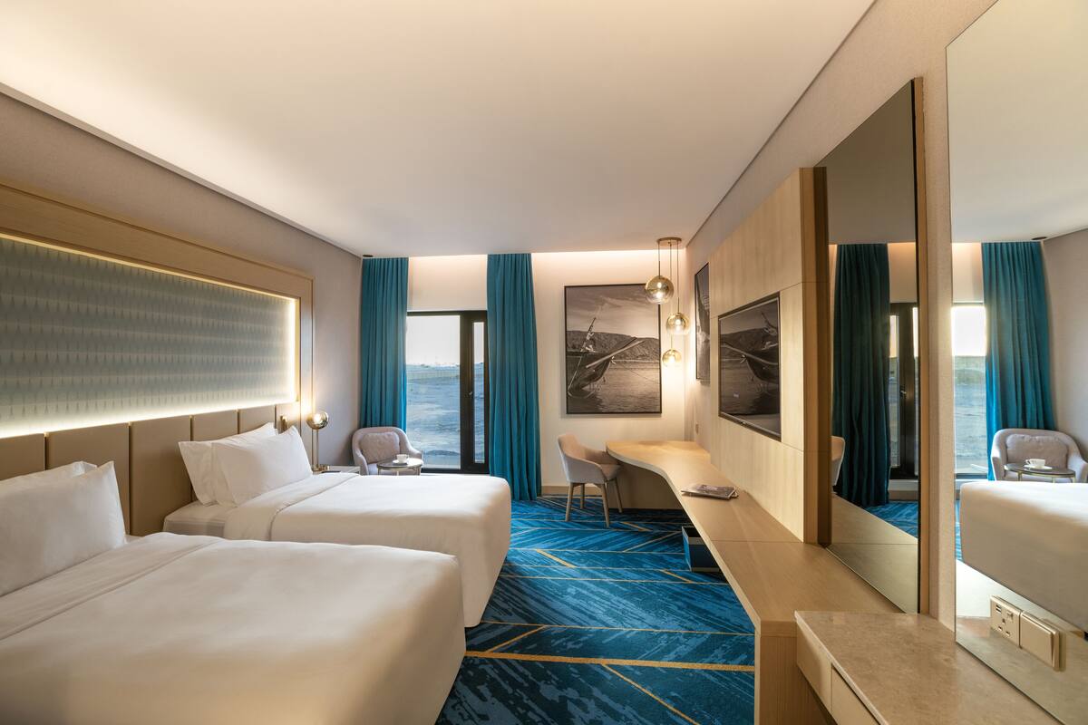 Une chambre d’hôtel avec deux lits, un bureau, une chaise, un grand miroir et une vue sur la mer.