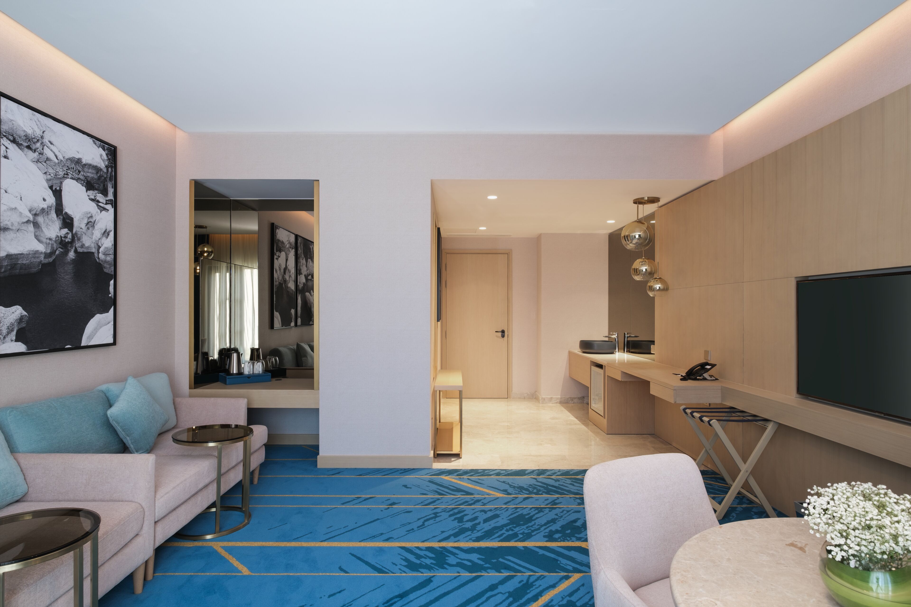 A modern hotel room with a living area featuring a sofa, a coffee table, and a TV. There is a kitchen area with a counter and a sink.