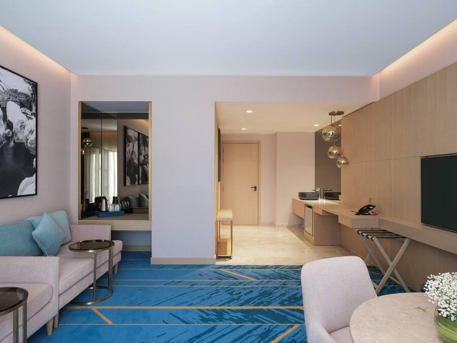 A modern hotel room with a living area featuring a sofa, a coffee table, and a TV. There is a kitchen area with a counter and a sink.