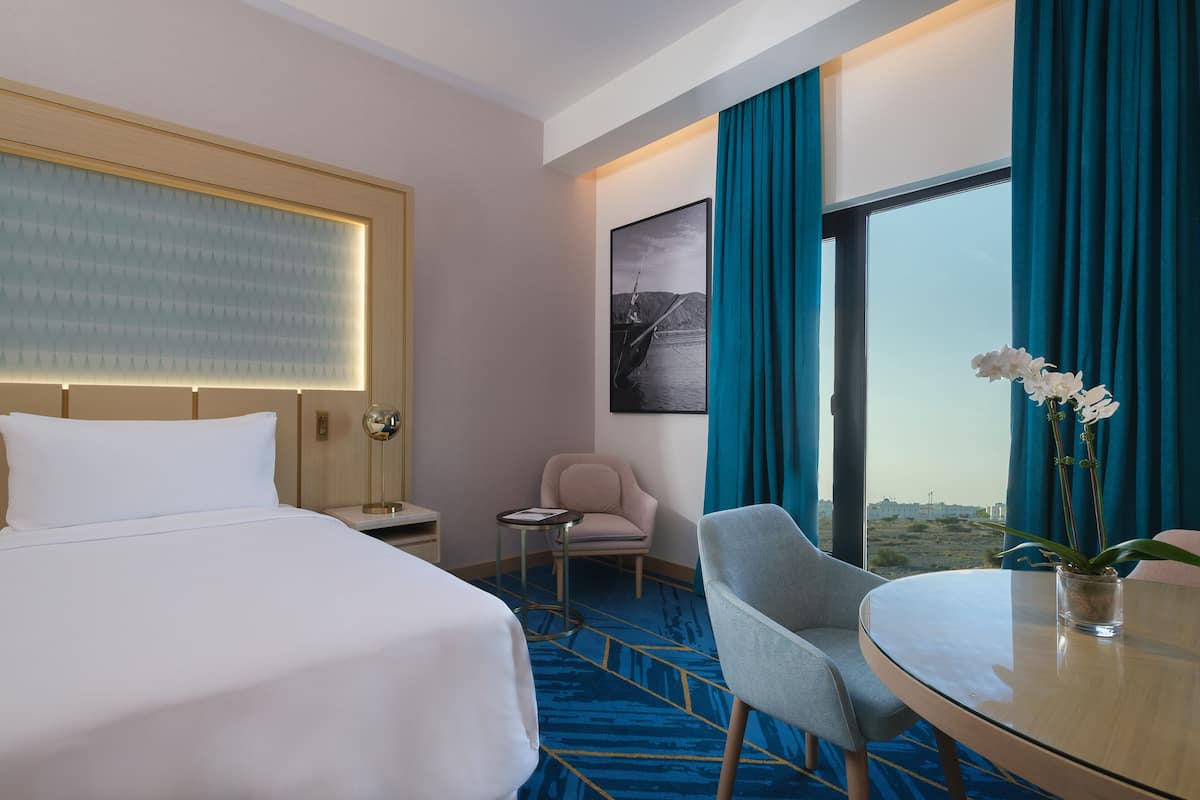 Une chambre d’hôtel avec un lit, une chaise, une petite table sur laquelle est posé un vase de fleurs, une table de chevet, une lampe, une fenêtre avec des rideaux et un tableau encadré au mur.