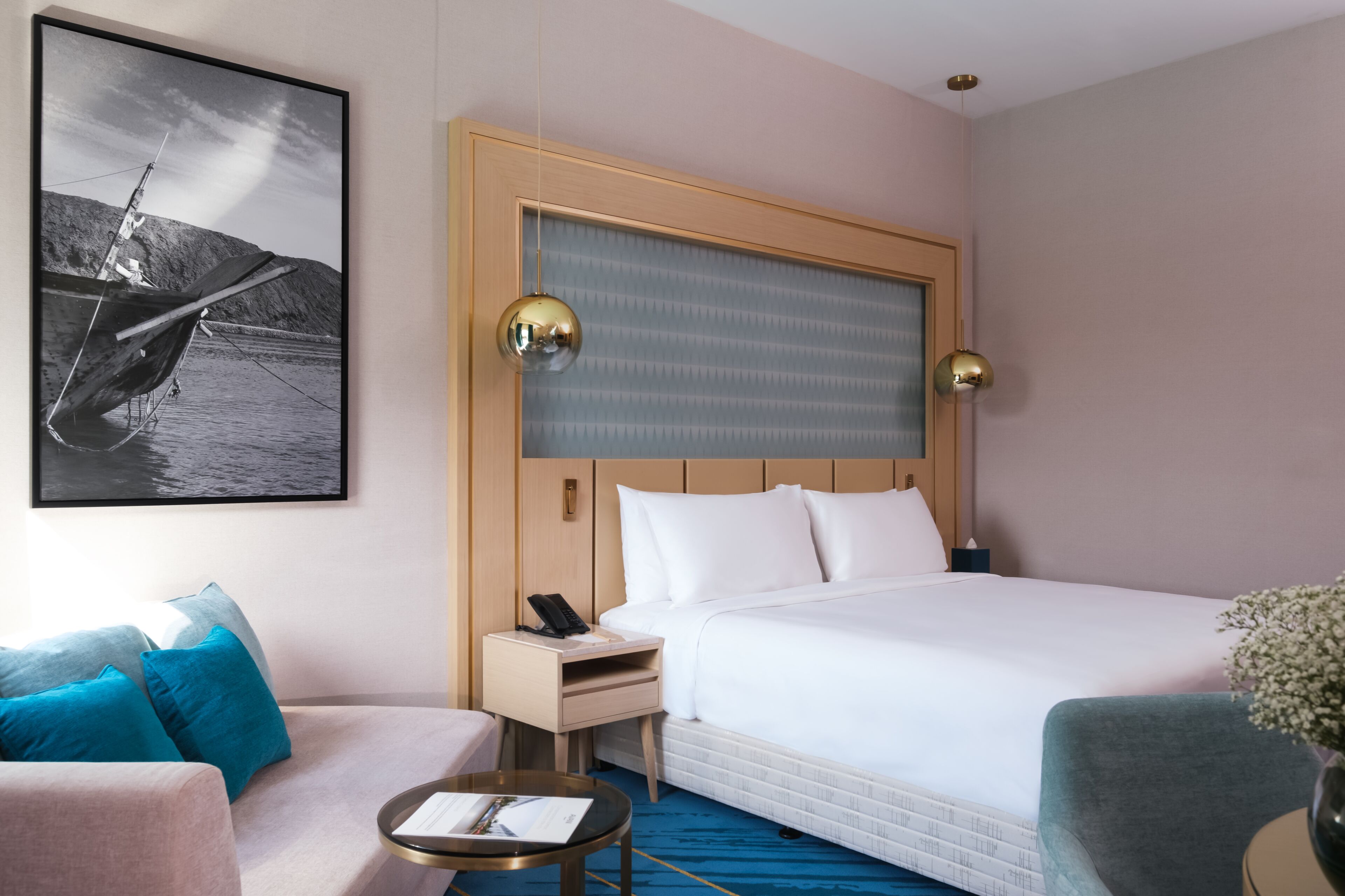 A hotel room with a large bed, a bedside table with a phone, a small table with a book, a sofa with blue cushions, and a framed picture on the wall.