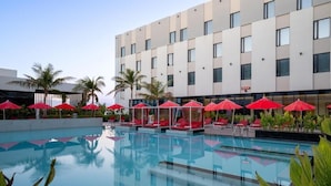 Um hotel moderno com piscina, guarda-sóis vermelhos e palmeiras.