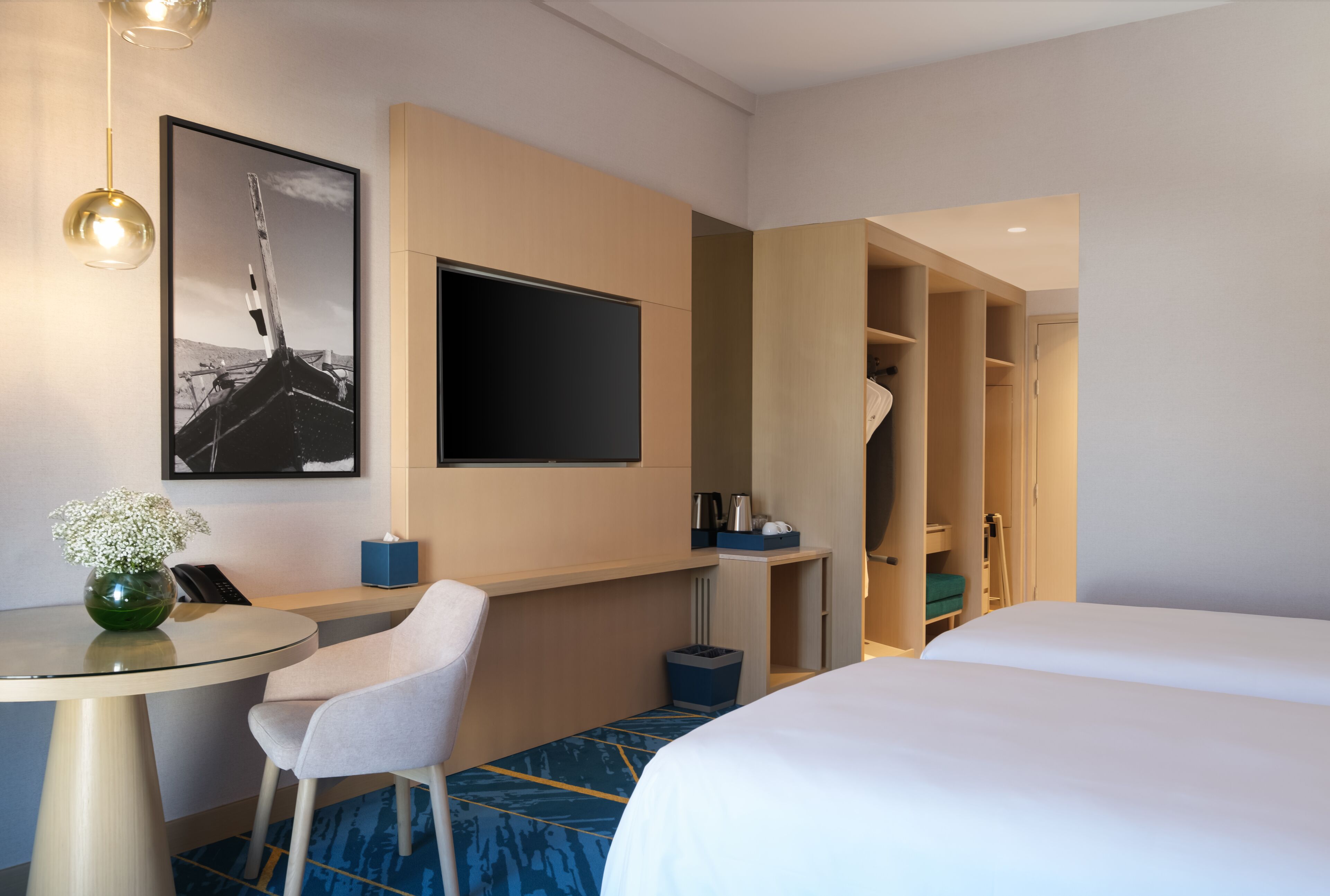 A hotel room with a bed, a desk with a chair, a small table with a vase, a coat closet, and a framed picture on the wall.