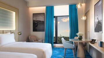 A hotel room with a large bed, a desk with a phone, a chair, a small table with a vase of flowers, and a view of the outdoors through a large window with blue curtains.