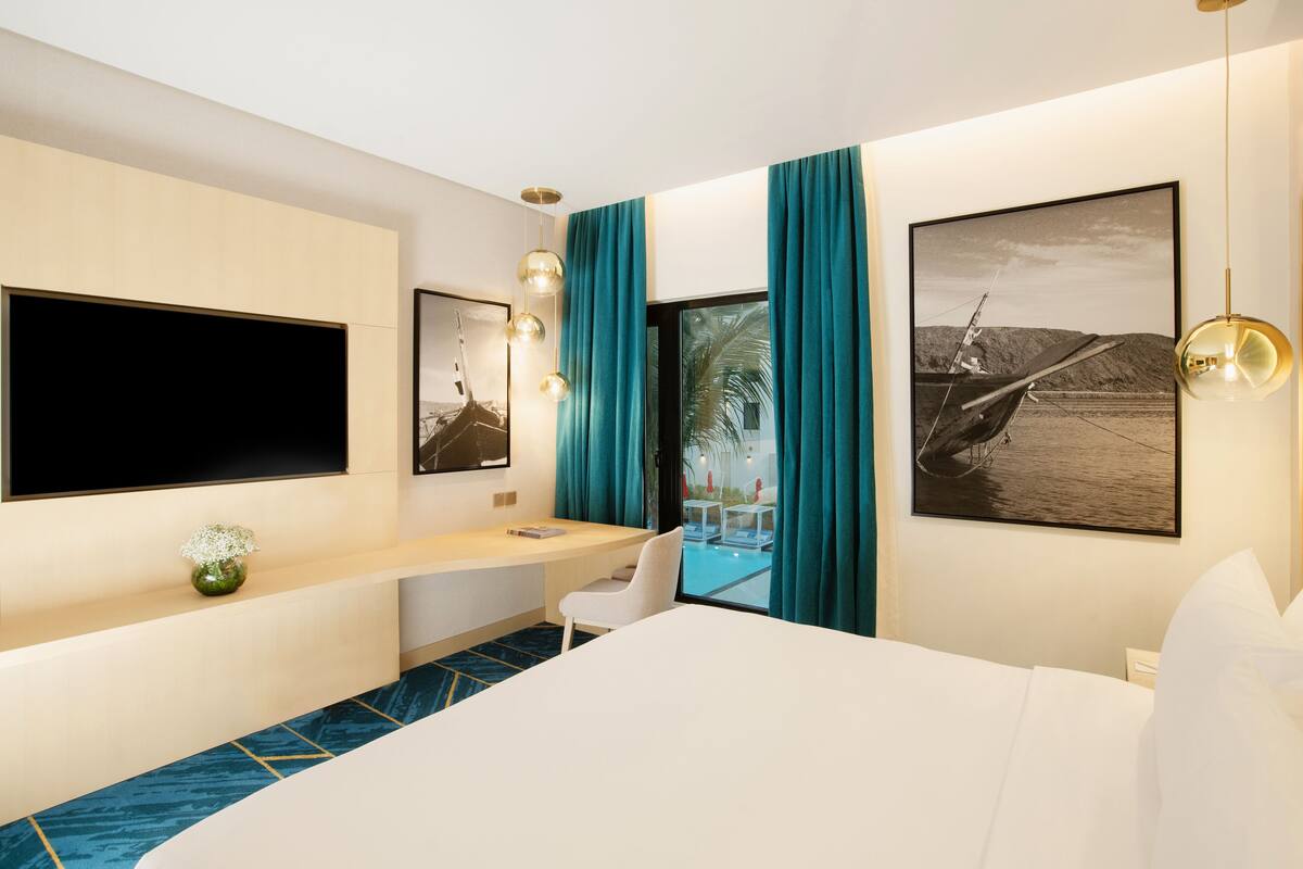 Une chambre d’hôtel moderne dotée d’un grand lit, d’un bureau avec une chaise, d’un téléviseur à écran plat et de deux grandes fenêtres ornées de rideaux turquoise.