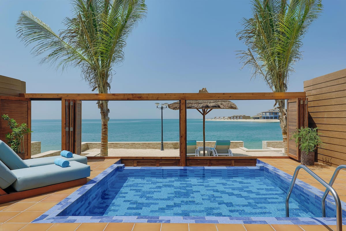 Un espace piscine avec vue sur la mer, une terrasse en bois, des chaises longues et un parasol en chaume.