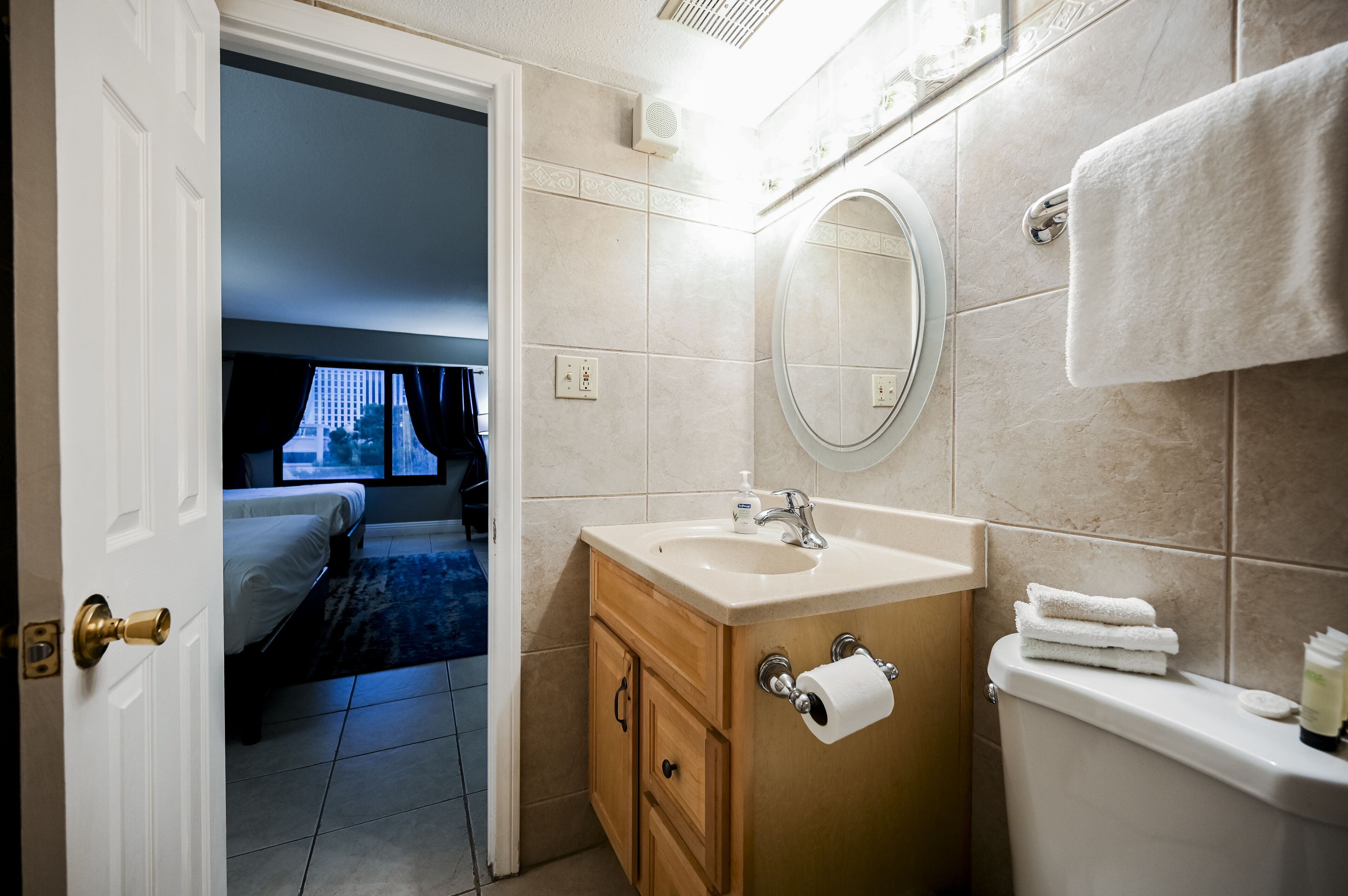 A hotel bathroom with a sink, mirror, and toilet. There is a door leading to a bedroom with a bed and a view of a building outside.