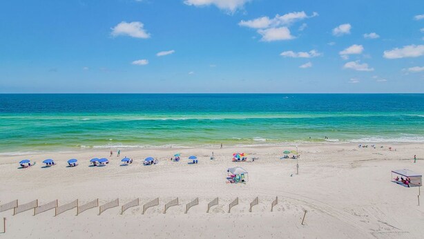 A beach with clear blue water, umbrellas, and a sandy shore. - Orange Beach Gulf-View 2BR • Sleeps 8 • Wet Bar • Kid-Friendly • Pools • Boats (Orange Beach)