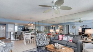 A spacious living room with a grey sofa, patterned armchairs, and a coffee table. A kitchen is visible in the background, and there is a large mirror reflecting the room. - Orange Beach Gulf-View 2BR • Sleeps 8 • Wet Bar • Kid-Friendly • Pools • Boats (Orange Beach)