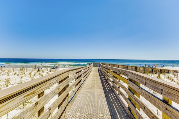 A wooden boardwalk leading to a beach with umbrellas and people.