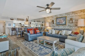 A spacious living room with a ceiling fan, two sofas, a coffee table, and a dining area with chairs and a kitchen in the background.