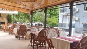 A covered outdoor dining area with tables and wicker chairs, a tiled floor, and a view of a building in the background. - Ozay Apart Hotel (Marmaris)