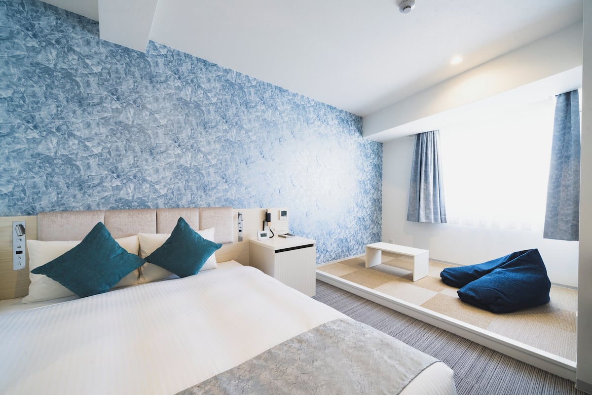 A modern hotel room with a large bed, a bedside table, a small desk, and a seating area with a bench and cushions.
