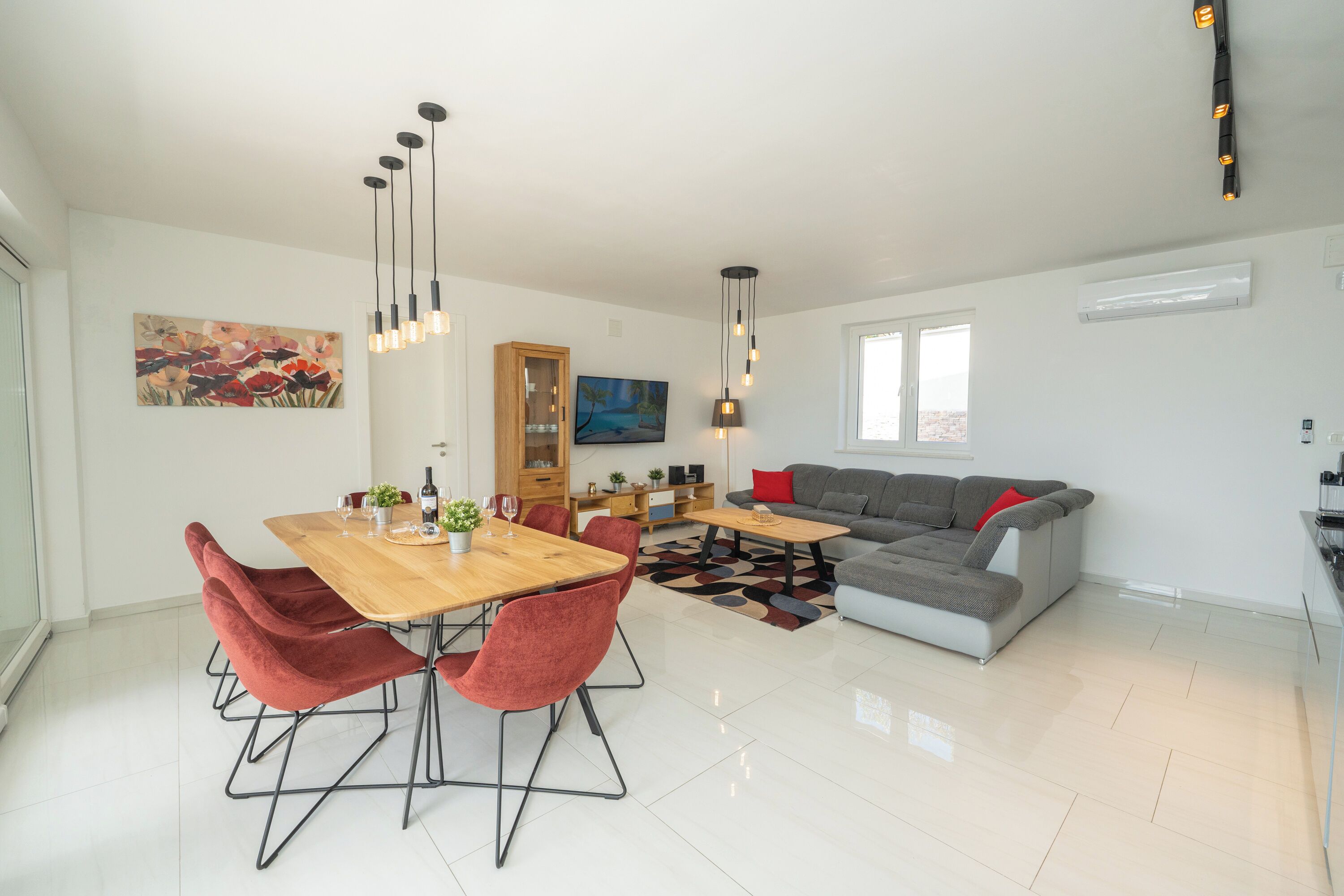 A modern living room with a dining area, a gray sofa, and a wooden dining table with red chairs.