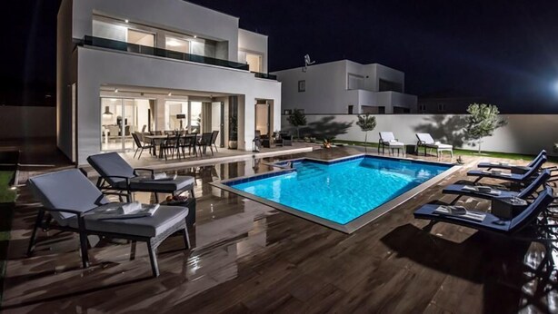 Ein moderner Poolbereich mit Liegestühlen, einem Schwimmbecken und einem zweistöckigen Gebäude mit offener Terrasse.