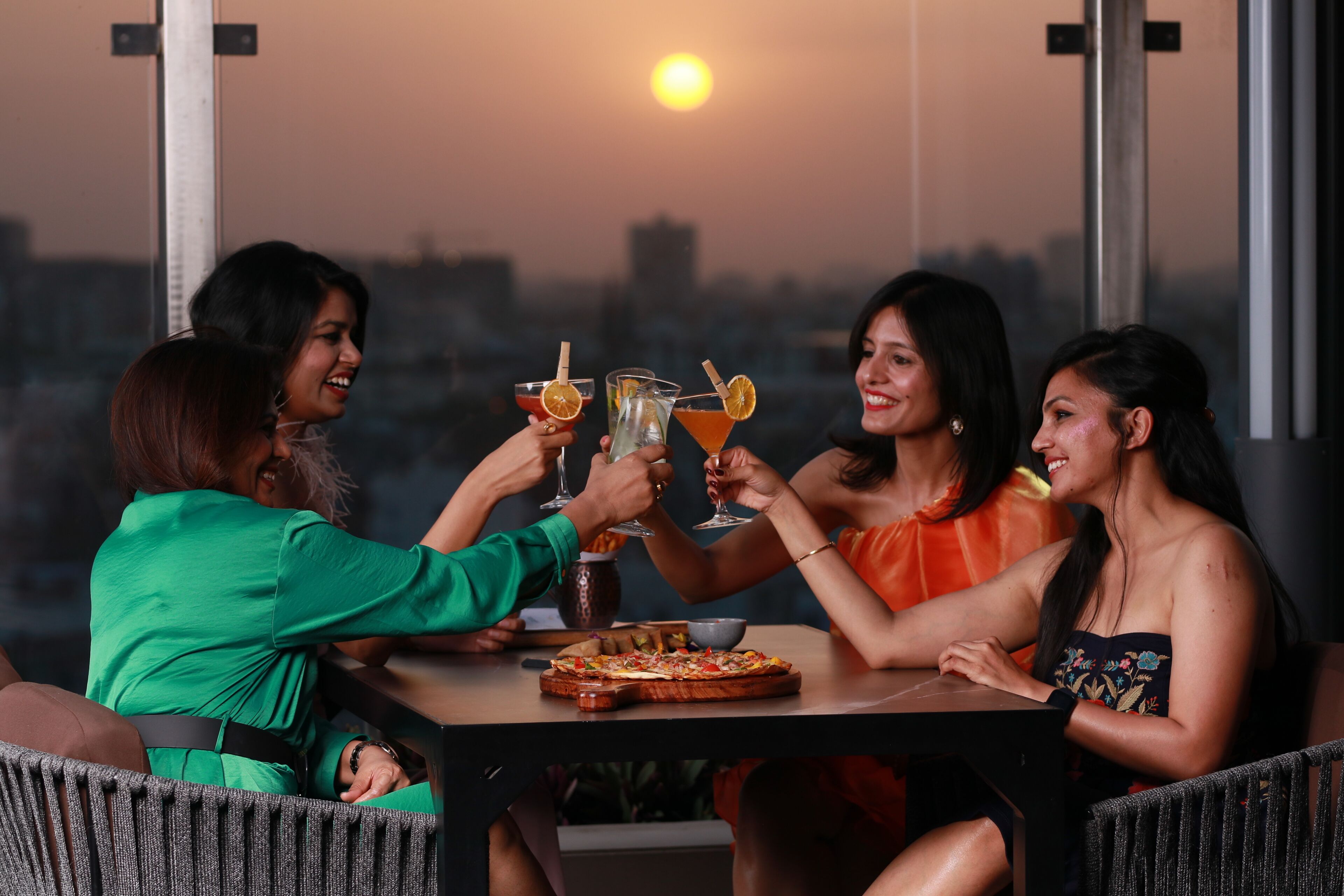 A group of people toasting with drinks at a table with a pizza, in a room with large windows and a city view.