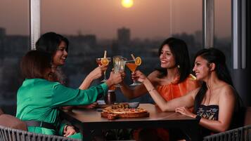 A group of people toasting with drinks at a table with a pizza, in a room with large windows and a city view.
