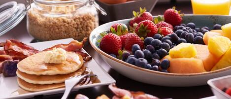 Una mesa de desayuno con panqueques, tocino, huevos y varias frutas.