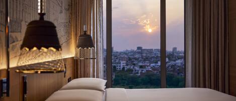 A hotel room with a large bed, a city view through the window, and a wall-mounted lamp.
