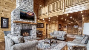 A spacious log cabin interior with a stone fireplace, multiple seating areas, and a balcony. - Serenity Mountain Pool Lodge, Theater Room, Sleeps 42, Indoor Pool!!! (Sevierville)