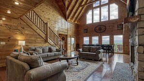 A spacious log cabin interior with a high ceiling, a staircase, a fireplace, and a large window overlooking a scenic view. - Serenity Mountain Pool Lodge, Theater Room, Sleeps 42, Indoor Pool!!! (Sevierville)
