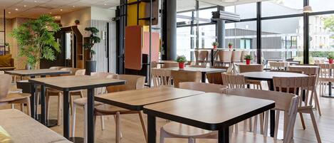 A modern cafe with wooden tables and chairs, large windows, and indoor plants.