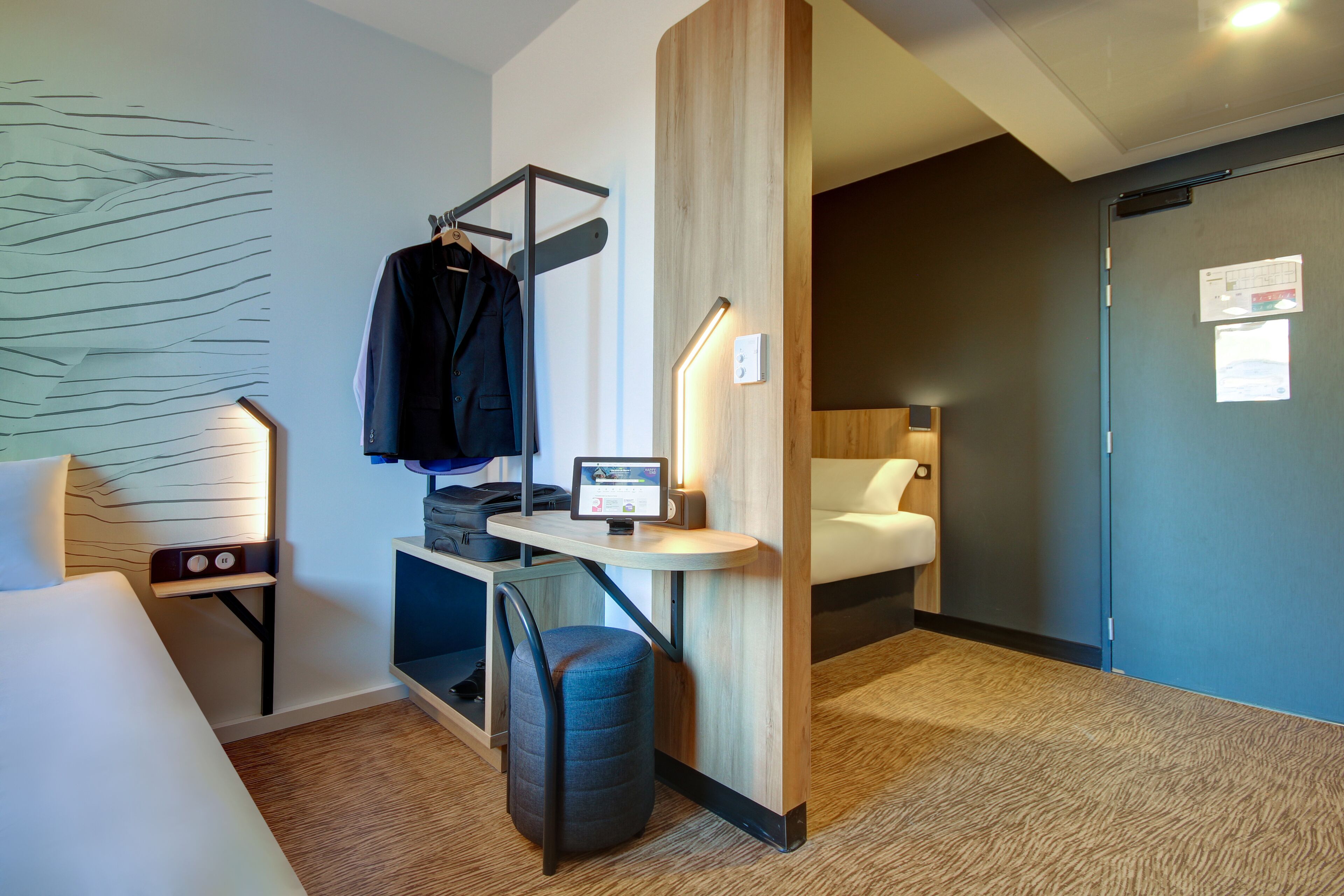 A hotel room with a bed, a desk with a computer, a chair, a coat rack, and a wall with a light fixture.