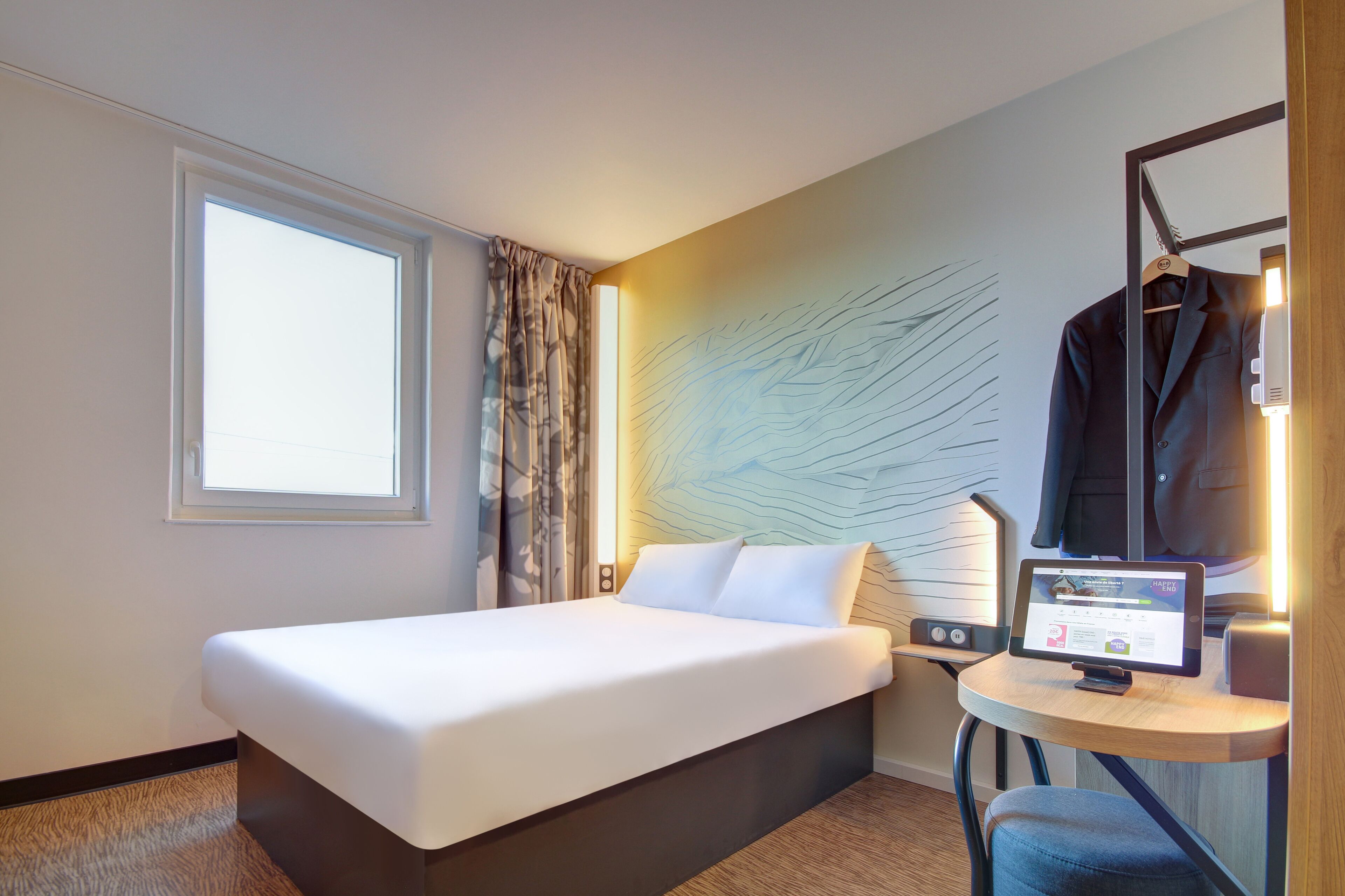 Ein Hotelzimmer mit einem Bett, einem Schreibtisch mit Computer, einem Stuhl, einem Kleiderhaken und einem Fenster mit Vorhängen.