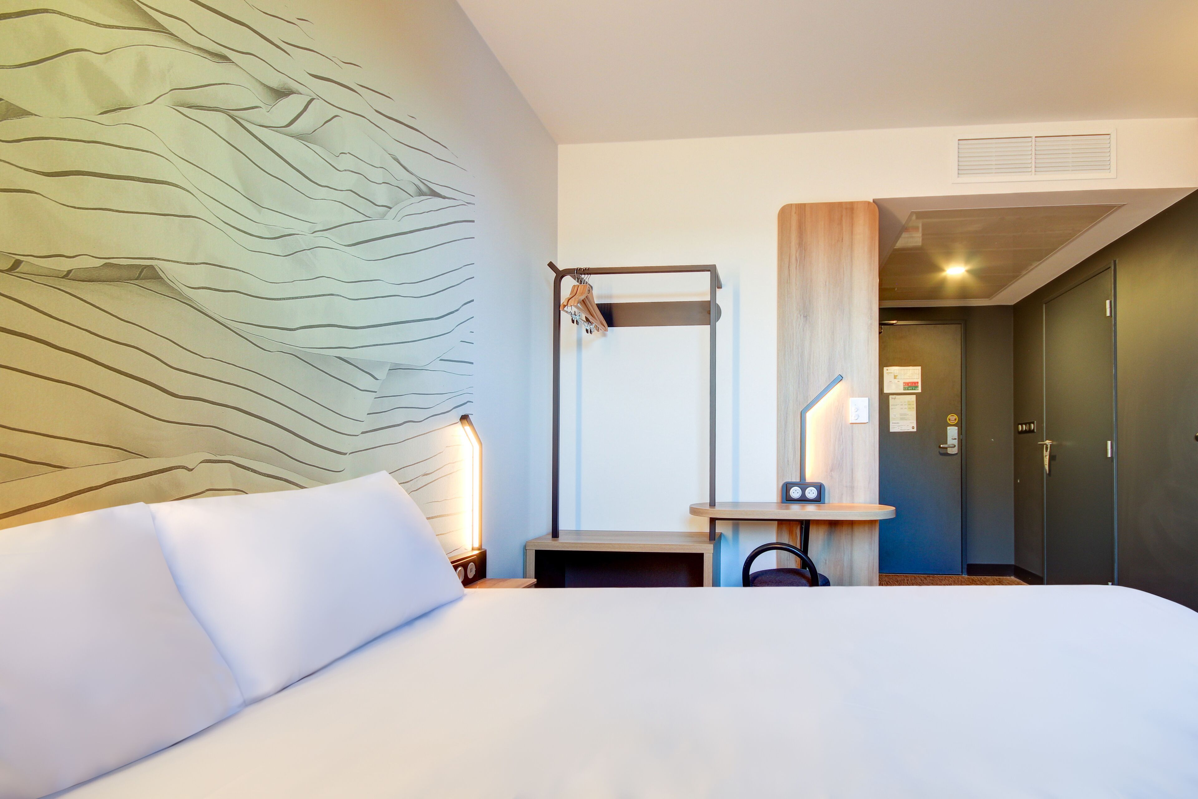 Habitación de hotel con cama, mesita de noche, espejo, luz en la pared y una puerta.