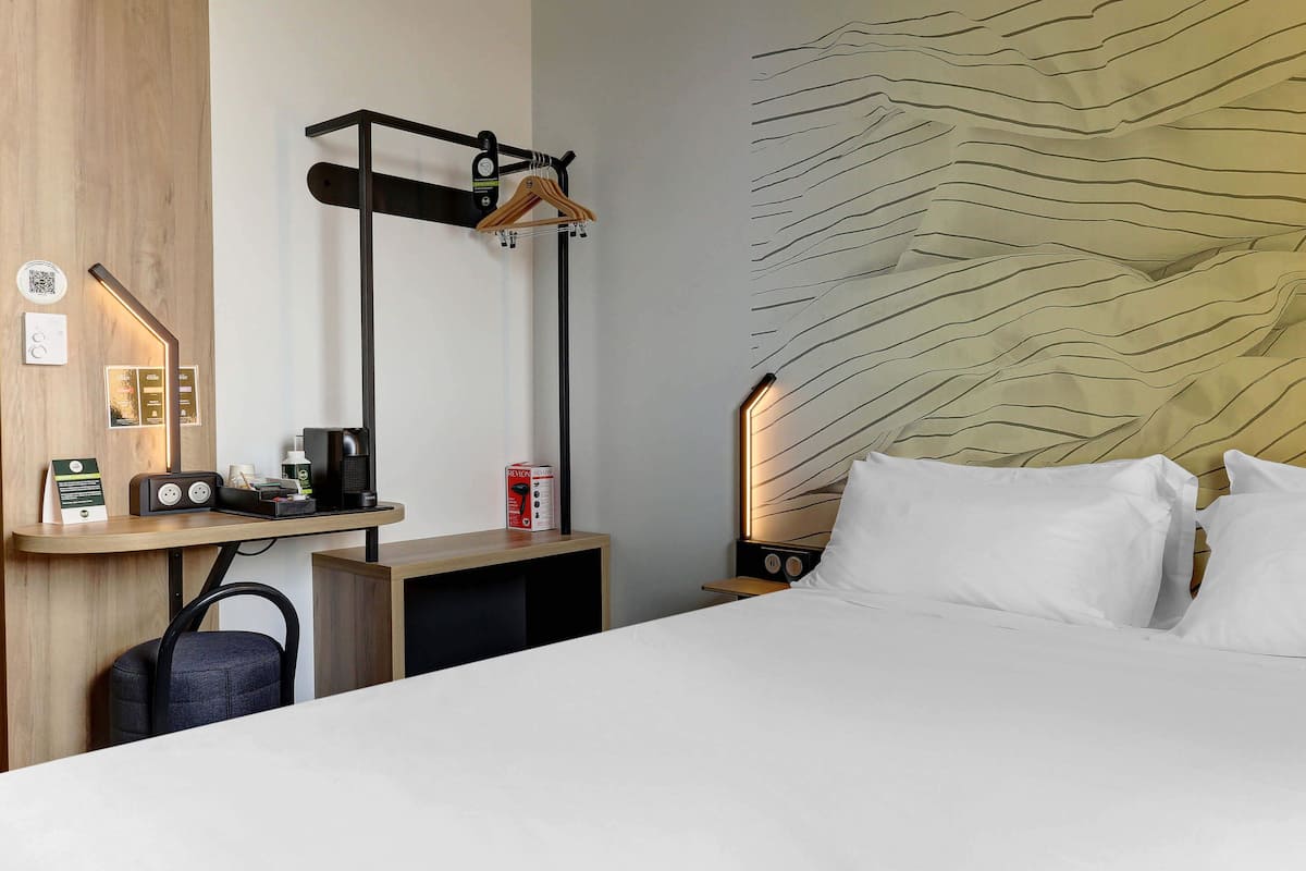 A hotel room with a bed, a desk with a coffee machine, a hanging coat rack, and a wall with a wavy pattern.