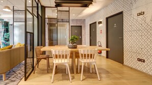 A modern dining area with a wooden table, white chairs, and a potted plant. There is a sofa with yellow cushions and a glass partition. - B&B HOTEL Epernay (Epernay)