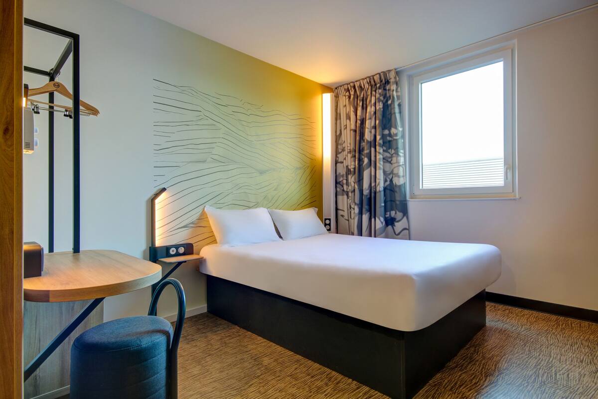 A hotel room with a bed, a desk, a chair, a bedside table, a lamp, a window with curtains, and a wall art piece.