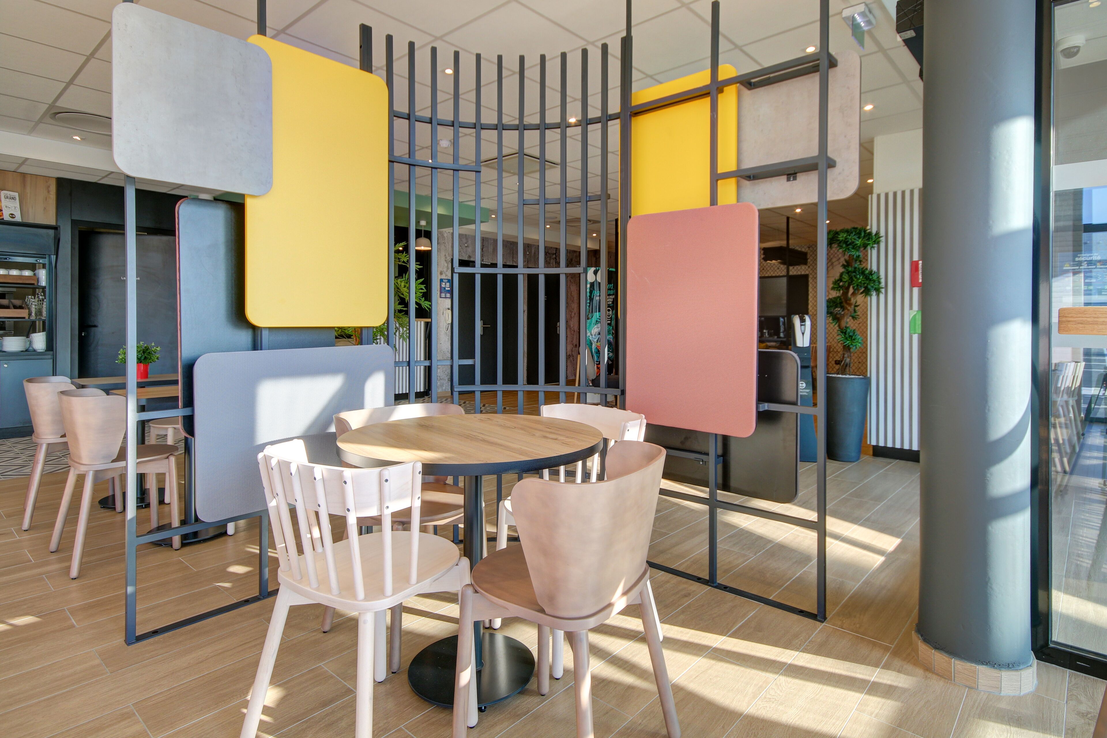 Spazio interno moderno con tavoli rotondi e sedie imbottite, un divisorio con barre verticali e vista all'esterno attraverso una porta in vetro.