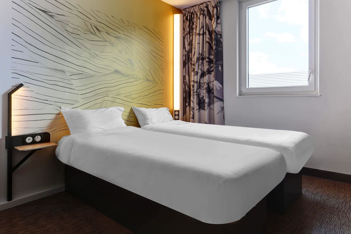 A double bed with white bedding, a nightstand with a lamp, and a large window with curtains.