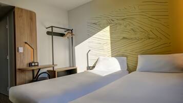 A hotel room with two beds, a wooden wardrobe, a desk with a chair, and a wall with a leaf pattern.